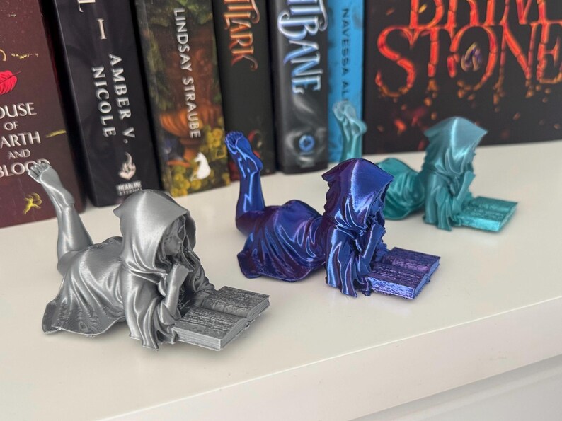 May include: Three figures of a hooded person reading a book. The figures are in silver, blue, and teal. The figures are lying down, with their legs extended. Books are in the background.
