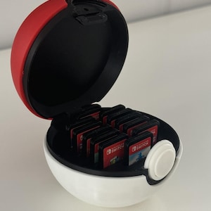 Könnte beinhalten: Eine weiße, rote und schwarze Pokéball-förmige Hülle, die geöffnet ist, um mehrere Nintendo Switch-Spielkassetten freizulegen. Die obere Hälfte ist rot, die Innenseite ist schwarz und die untere Hälfte ist weiß mit einem weißen Knopfdetail.