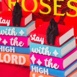 May include: Two stacks of miniature books in red, blue, pink, and orange with white edges. Each stack is topped with a small black hooded figure. White raised lettering on the spines reads "stay with the HIGH LORD" with small star icons.