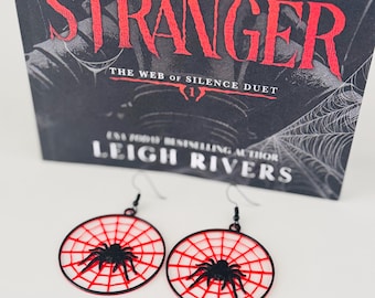 May include: Spiderweb earrings with a black spider design and red web detail. The earrings are round and hang from black hooks. A book titled "Little Stranger" is in the background.