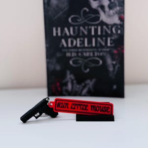 May include: A black handgun keychain with a red tag that reads "RUN LITTLE MOUSE" is displayed in front of a book titled "HAUNTING ADELINE". The book cover is black with white text and a skull graphic.