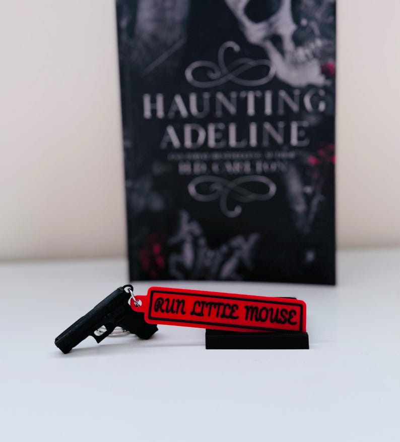 May include: A black handgun keychain with a red tag that reads "RUN LITTLE MOUSE" is displayed in front of a book titled "HAUNTING ADELINE". The book cover is black with white text and a skull graphic.