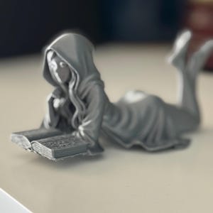 May include: A grey figurine of a person in a hooded robe, reclining and reading a book. The figure is lying down, with the book open in front of them. The figurine is a solid grey colour and appears to be made of a hard material.
