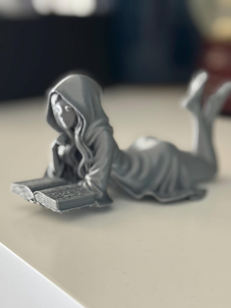 May include: A grey figurine of a person in a hooded robe, reclining and reading a book. The figure is lying down, with the book open in front of them. The figurine is a solid grey colour and appears to be made of a hard material.
