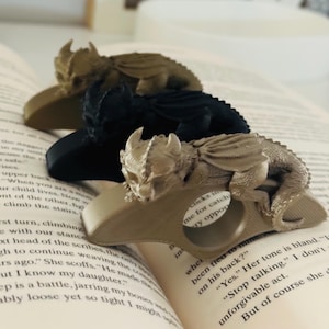 May include: Three dragon-shaped book rings in varying colors: gold, black, and bronze. Each dragon rests on a ring base, designed to hold a book open. The rings are displayed on the pages of an open book.