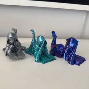 May include: Three figurines of cloaked figures in silver, teal, and blue, each reclining and reading a book. The figures are detailed with flowing robes and hoods, and the books have visible text. The figurines are on a white surface.