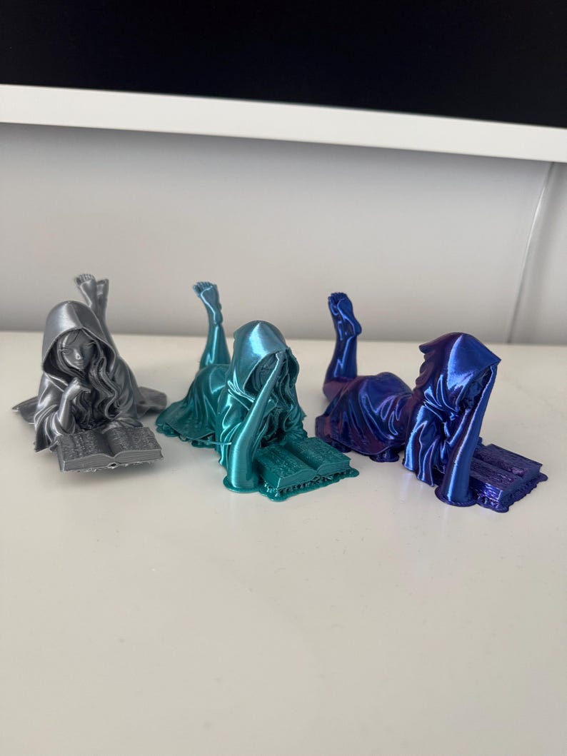 May include: Three figurines of cloaked figures in silver, teal, and blue, each reclining and reading a book. The figures are detailed with flowing robes and hoods, and the books have visible text. The figurines are on a white surface.