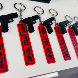 May include: A collection of black gun-shaped keychains with silver key rings. Each keychain has a red rectangular tag with the words "RUN LITTLE MOUSE" in black lettering.