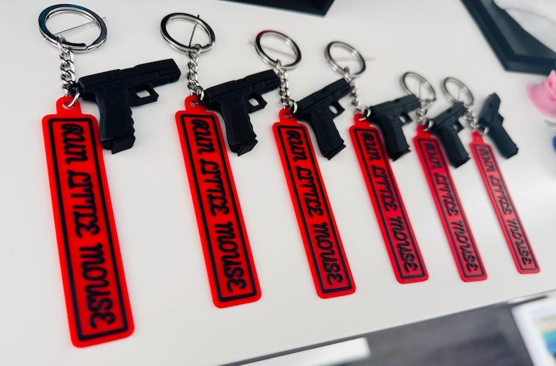 May include: A collection of black gun-shaped keychains with silver key rings. Each keychain has a red rectangular tag with the words "RUN LITTLE MOUSE" in black lettering.