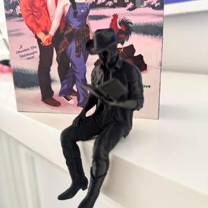 May include: A black figurine of a person in a cowboy hat and boots, holding a book, seated on a white surface. The figurine is positioned before a book cover featuring a couple and the text "A Mountain Men Matchmaker novel".