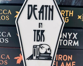 May include: A white coffin-shaped sticker with black trim and the words "DEATH BY TBR" in a gothic font. Below the text is a skull resting on books. The sticker is placed in front of a stack of books.