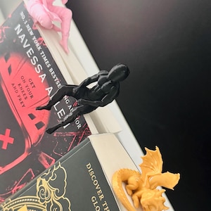 May include: Three unique book page holders. A pink figure sits atop a book, a black figure reclines on another, and a golden dragon rests on a third. The books have titles and author names visible.