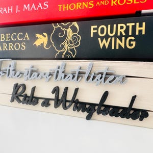 May include: A selection of books with the titles "Thorns and Roses" and "Fourth Wing" visible. Below the books are two decorative phrases: "To the stars that listen" in grey and "Ride a Wingleader" in black.