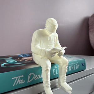 May include: A white 3D printed figurine of a hockey player wearing a jersey, helmet, and skates, sitting on the edge of a book titled The Deal by Elle Kennedy. The figure is holding a small open book in its hands while in a seated position.