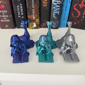 May include: Three bookends shaped as figures reading books. The bookends are in blue, teal, and silver. They are on a white shelf in front of a row of books.