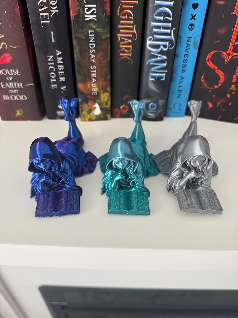 May include: Three bookends shaped as figures reading books. The bookends are in blue, teal, and silver. They are on a white shelf in front of a row of books.