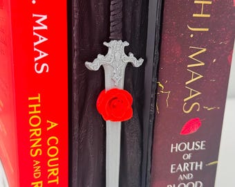 May include: A silver sword-shaped bookmark with a red rose accent, placed between two books by Sarah J. Maas. The books have red and burgundy covers with white text. The bookmark is in a black case.