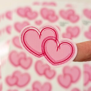 May include: Two pink heart stickers with a red brushstroke outline are held up against a background of other heart stickers. The stickers are a light pink color with a darker pink outline. The image is well-lit and the stickers are in focus.