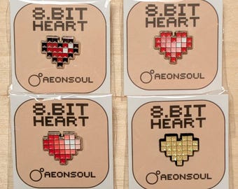 Puede incluir: Cuatro pines de esmalte con diseños de corazón pixelados en rojo, rosa y dorado. Cada pin está en una tarjeta de respaldo beige con el texto "8.BIT HEART" y el nombre de la marca "AEONSOUL". Los pines están empaquetados en plástico transparente.