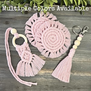 May include: A collection of pink macrame accessories, including coasters, a car air freshener, and a keychain with wooden beads. The text "Multiple Colors Available" is at the top of the image. The items are arranged on a wooden surface.