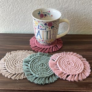May include: A white ceramic mug with the words "have faith" and floral designs sits on a pink macrame coaster. Three additional macrame coasters in beige, green, and pink are also visible. The coasters have a spiral design with fringe.
