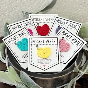 May include: A collection of crochet heart charms in various colors, each attached to a small card. The cards are white with the words "POCKET VERSE" printed on them. The yellow heart card has a Bible verse printed on it.