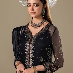Puede incluir: Una mujer con un vestido formal negro con adornos dorados y una capa transparente. El vestido tiene un escote en V y está adornado con lentejuelas. Lleva joyas a juego, incluyendo un collar, pendientes, pulseras y anillos.