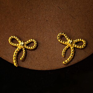May include: A pair of gold-coloured bow-shaped stud earrings. The earrings are small and detailed, with a textured surface. They are set against a brown background.