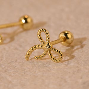May include: A pair of gold-coloured bow-shaped stud earrings. The bows have a textured, rope-like design. Each earring has a small, round ball on the back for securing the earring. The earrings are displayed on a light-coloured surface.