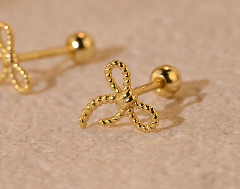 May include: A pair of gold-coloured bow-shaped stud earrings. The bows have a textured, rope-like design. Each earring has a small, round ball on the back for securing the earring. The earrings are displayed on a light-coloured surface.