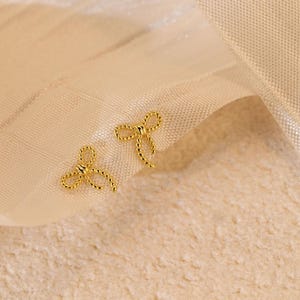 May include: A pair of gold-coloured bow-shaped stud earrings. The earrings are displayed on a cream-coloured fabric background. The bows have a textured, twisted rope design.