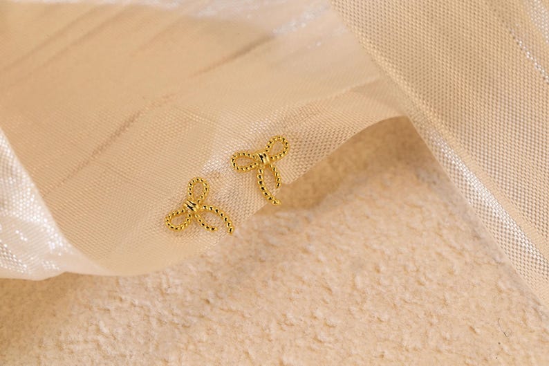 May include: A pair of gold-coloured bow-shaped stud earrings. The earrings are displayed on a cream-coloured fabric background. The bows have a textured, twisted rope design.
