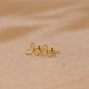 May include: A pair of gold-coloured bow-shaped stud earrings. The earrings have a textured, rope-like design and a small, round post. The earrings are displayed on a neutral, light brown surface.