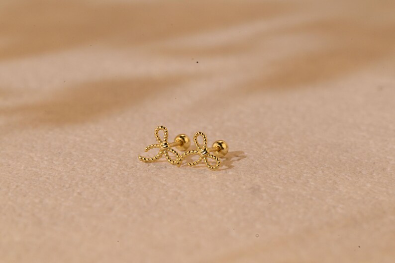 May include: A pair of gold-coloured bow-shaped stud earrings. The earrings have a textured, rope-like design and a small, round post. The earrings are displayed on a neutral, light brown surface.