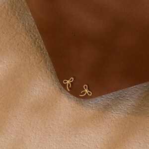 May include: A pair of small, gold-coloured bow-shaped stud earrings. The earrings are positioned on a brown surface, with a soft, light-coloured background. The bows have a delicate, detailed design.