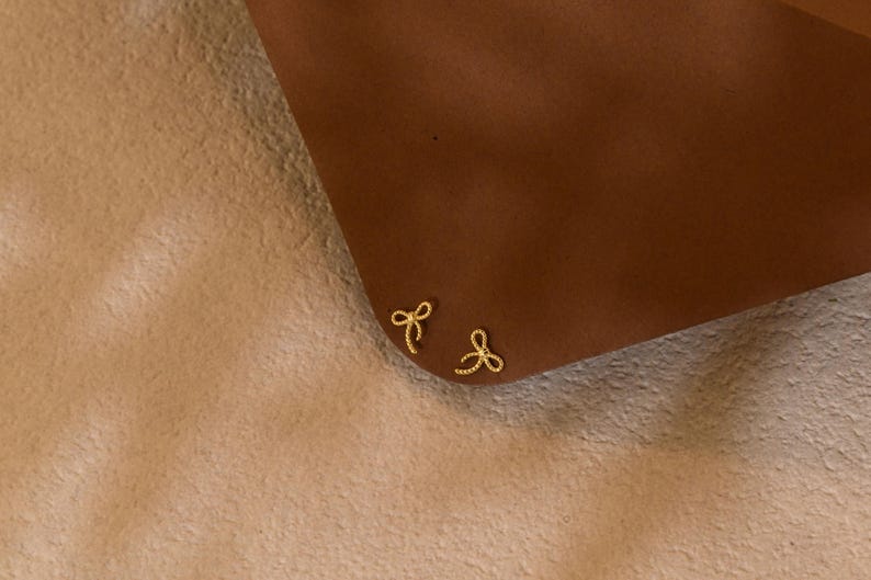 May include: A pair of small, gold-coloured bow-shaped stud earrings. The earrings are positioned on a brown surface, with a soft, light-coloured background. The bows have a delicate, detailed design.