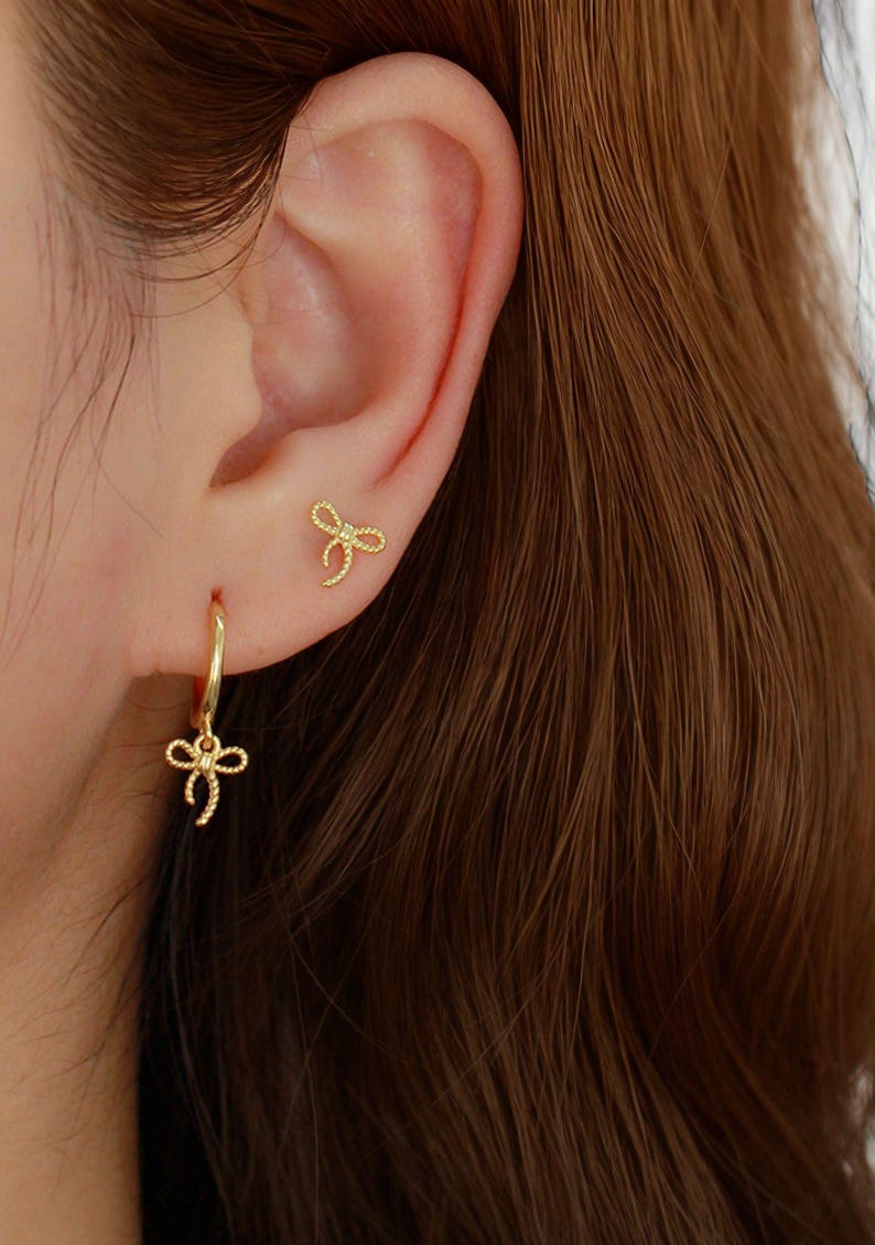 May include: Gold-coloured bow earrings. One is a small stud, the other a hoop earring with a bow charm. The earrings are on a person's ear, with brown hair in the background. The bow design is a delicate, feminine style, suitable for everyday wear.