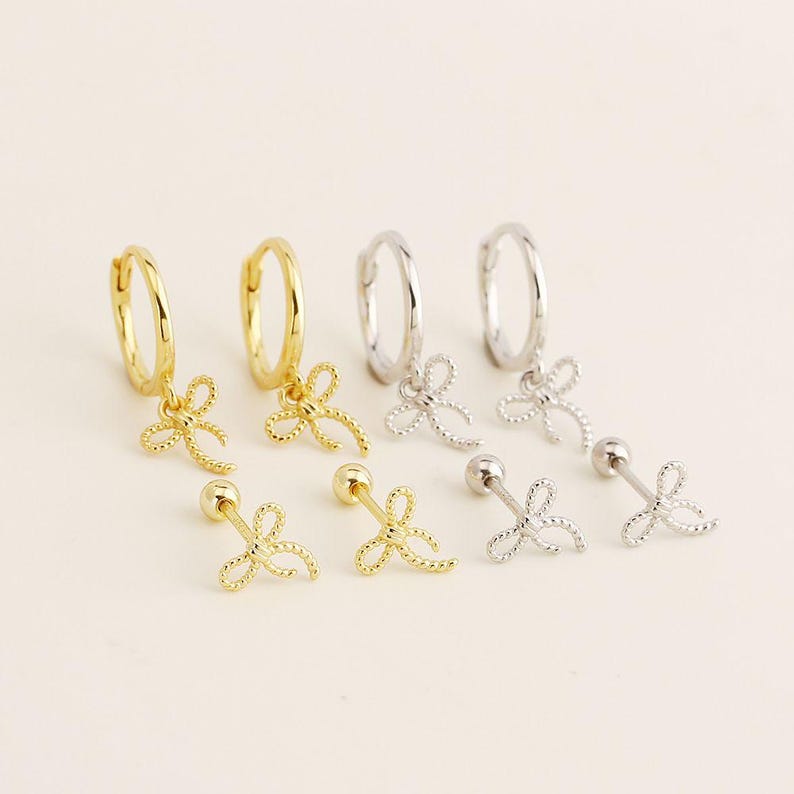 May include: A selection of gold and silver earrings. The image shows small hoop earrings with bow charms, and stud earrings with bow designs. The earrings are presented on a light-coloured surface.