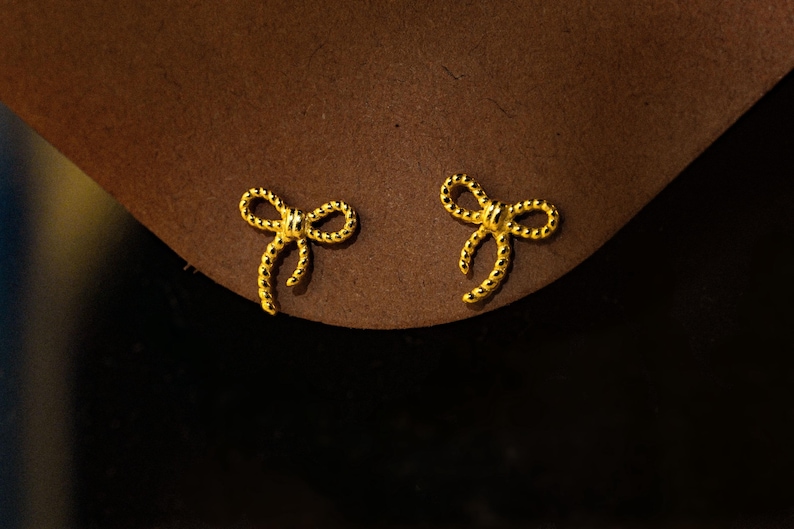 May include: A pair of gold-coloured bow-shaped stud earrings. The earrings are small and detailed, with a textured surface. They are set against a brown background.