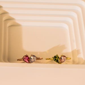 May include: Two rings with gemstone accents. One ring has a rose gold band with a pink and clear gemstone design. The other ring has a gold band with green and pink gemstones. Both rings have a teardrop shape.
