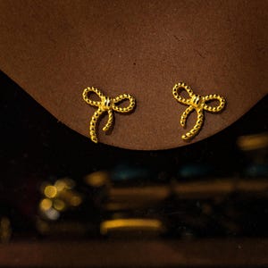 May include: Two gold-coloured bow-shaped stud earrings. The earrings are set against a brown surface, with a dark background. The bows have a textured finish, and the overall look is elegant and delicate.