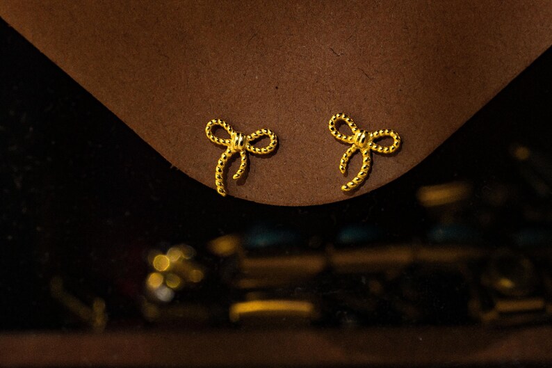 May include: Two gold-coloured bow-shaped stud earrings. The earrings are set against a brown surface, with a dark background. The bows have a textured finish, and the overall look is elegant and delicate.