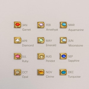 Puede incluir: Una colección de colgantes dorados, cada uno con una piedra preciosa y un mes del año. Las piedras preciosas incluyen granate, amatista, aguamarina, diamante, esmeralda, piedra lunar, rubí, peridoto, zafiro, ópalo, citrino y turquesa.
