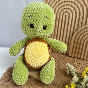 May include: A plush turtle toy crafted from soft, light green yarn with a textured, velvety appearance. The turtle features a round, yellow belly patch framed by a brown border, dark brown plastic eyes, and stitched eyebrows on its head.