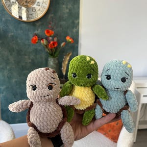 May include: Three small, handmade crochet turtle plushies featuring soft, textured yarn. One turtle is light beige, one is vibrant green with a yellow belly, and one is light blue. Each has large black eyes and small, detailed shell markings.