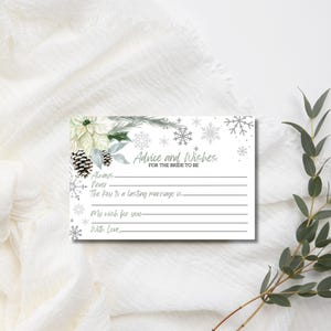 May include: A white card with the text "Advice and Wishes for the Bride to Be" and lines for writing. The card features a winter theme with snowflakes, pinecones, and floral accents in white and silver.
