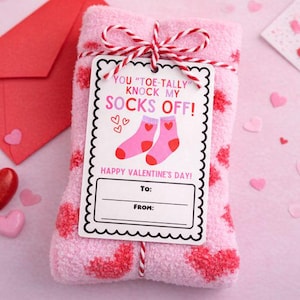 May include: A pink Valentine's gift set with socks patterned with red hearts, tied with red and white string. A tag reads "YOU 'TOE-TALLY' KNOCK MY SOCKS OFF!" with "HAPPY VALENTINE'S DAY!" below. A red envelope and heart-shaped sweets are also present.
