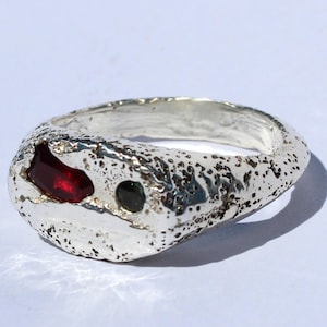Peut inclure: Une bague en argent texturé au design unique. La bague présente un large anneau de forme irrégulière avec une surface rugueuse et piqueté. Elle comporte une pierre précieuse rouge foncé et une petite pierre précieuse noire.