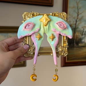 May include: A decorative wall hanging featuring a pastel mint and pink moth sculpture with realistic eyelashes on the wings, mounted on a small ornate gold frame. The moth is adorned with gold star accents and two dangling gold crescent moon charms.