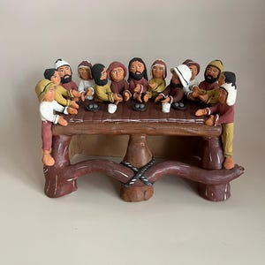 Puede incluir: Una escultura artesanal de madera marrón que representa a un grupo de figuras sentadas alrededor de una mesa. Las figuras visten ropa de varios colores, incluyendo amarillo, rojo y marrón. La mesa tiene una base curva única con un detalle de cuerda.